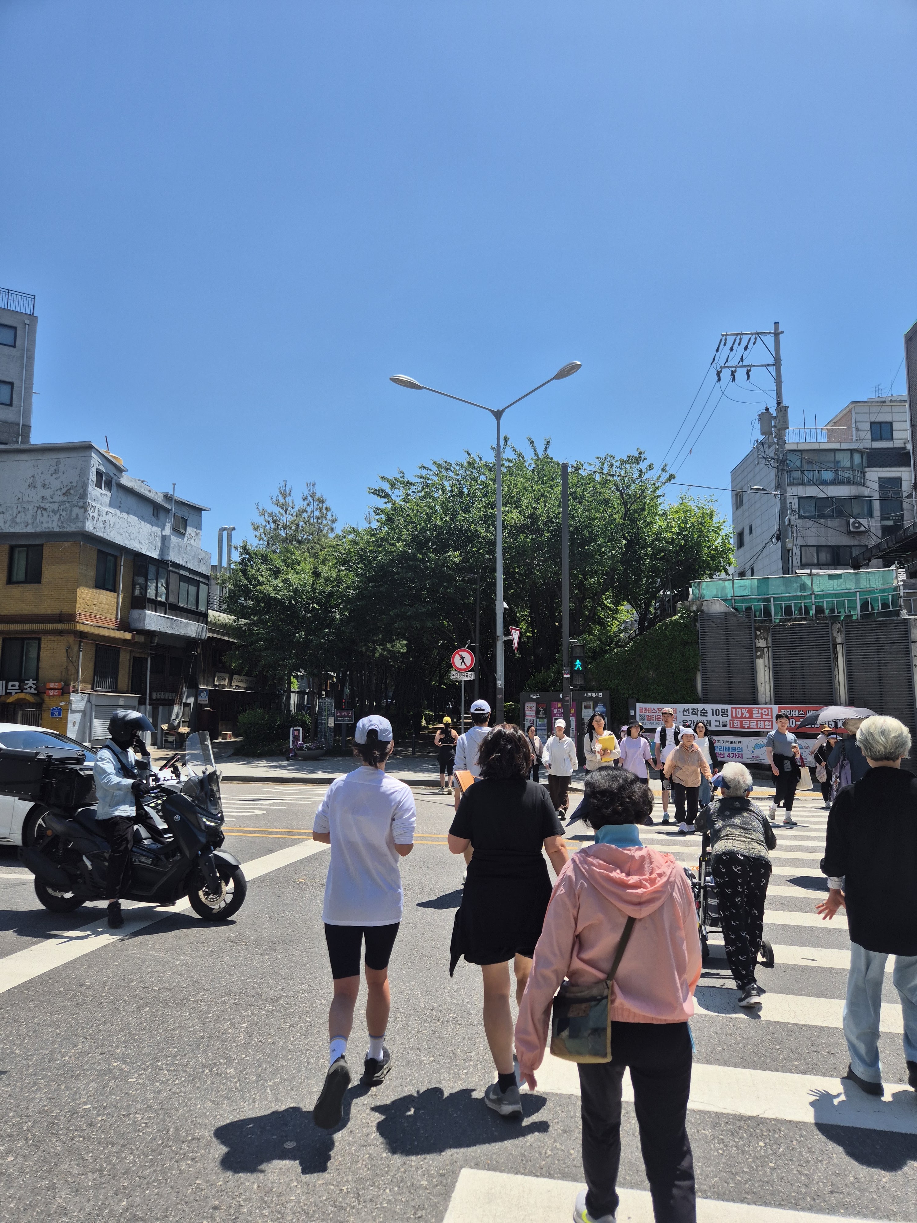 A busy street scene with pedestrians crossing at a zebra crossing on a sunny day. The group includes people of various ages, with a mix of casual and sporty clothing. A motorcycle is visible in the foreground, alongside buildings and green trees in the background.