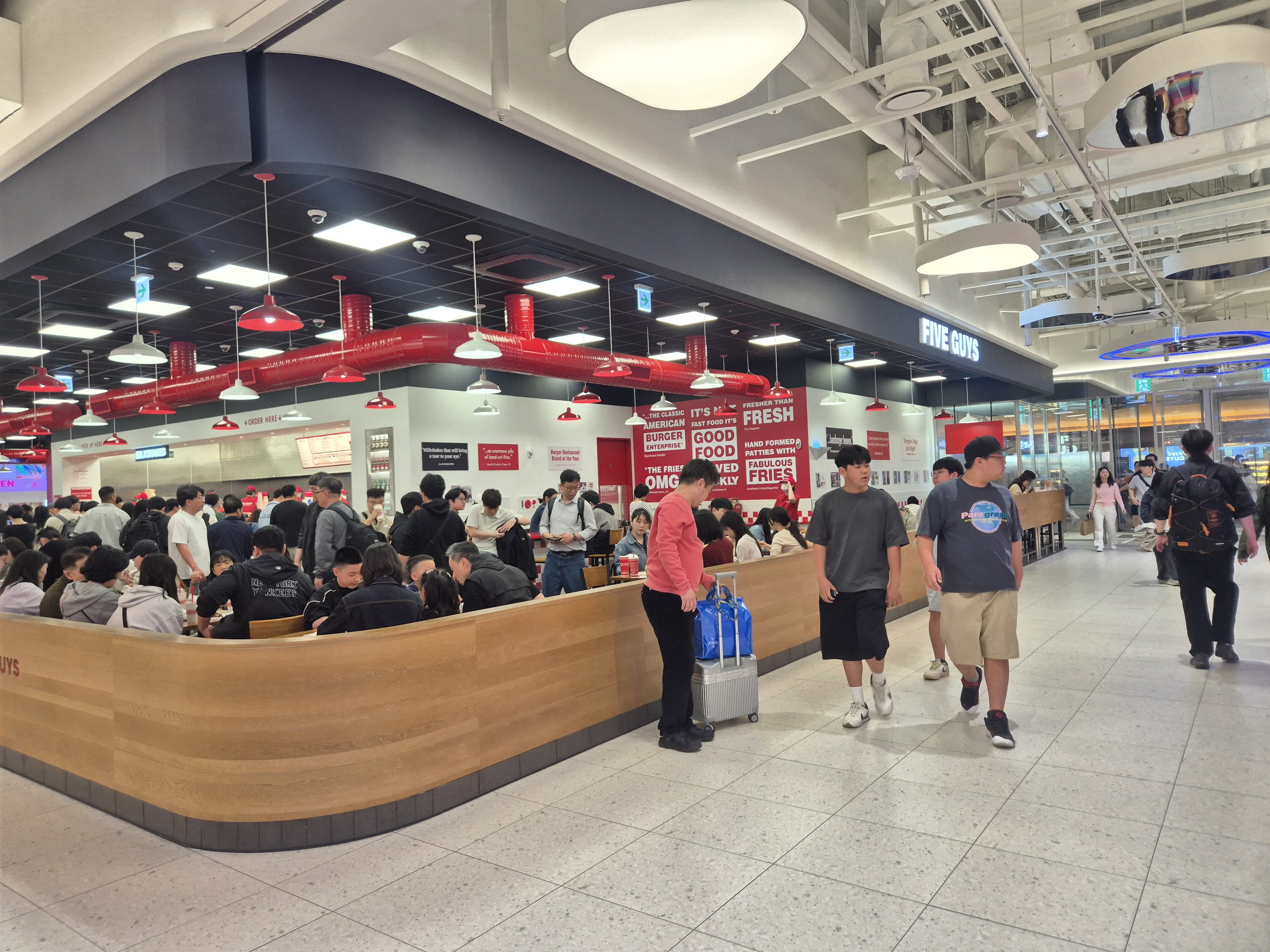 A busy indoor food court featuring a Five Guys restaurant, with many customers seated and a queue at the counter. The decor includes red lighting and vibrant signage promoting the restaurant's offerings.