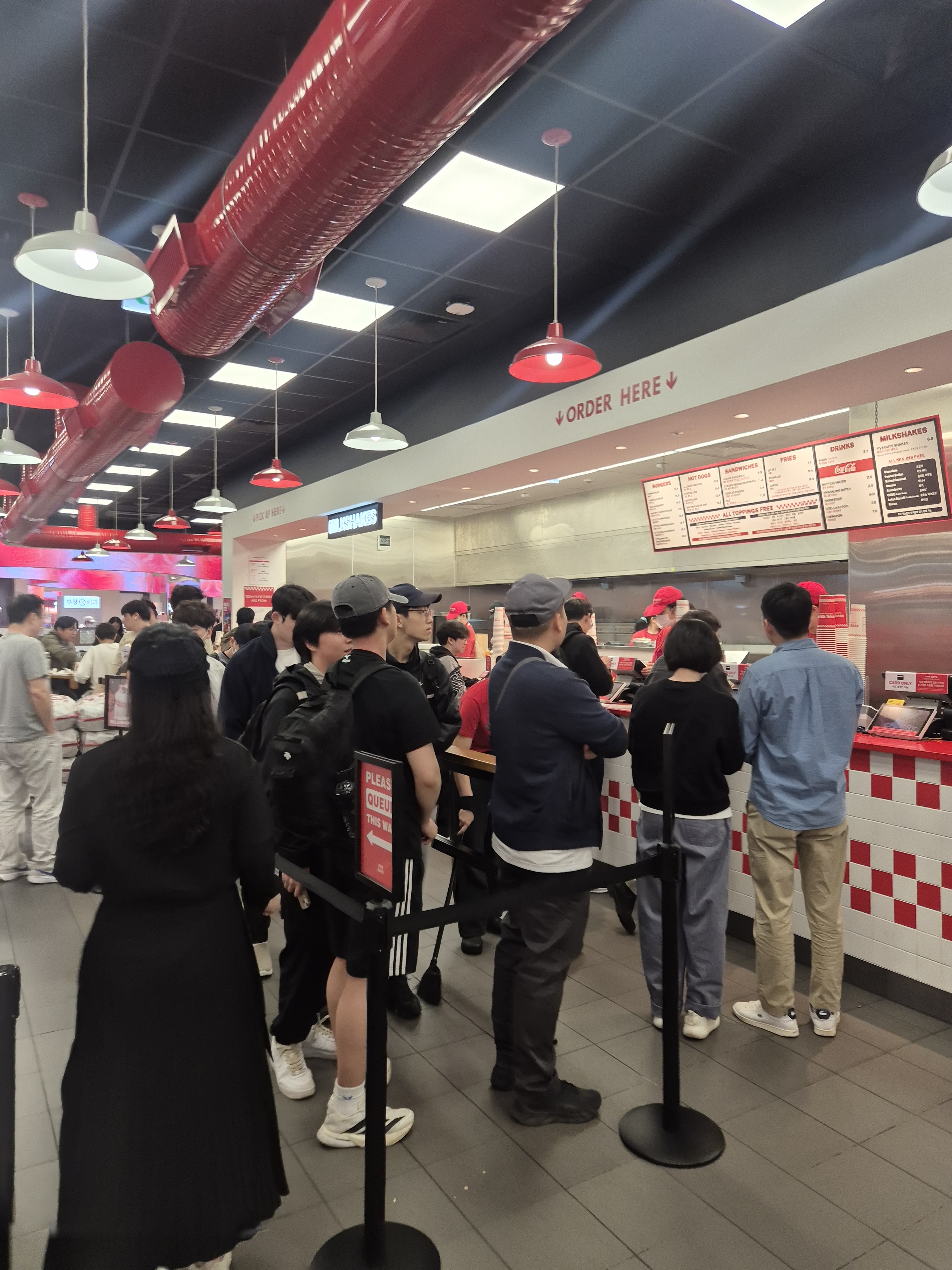 A busy restaurant interior with patrons queuing at the counter, which displays an 'Order Here' sign. The decor features red and white colours, with hanging lights and a visible menu board behind the staff.