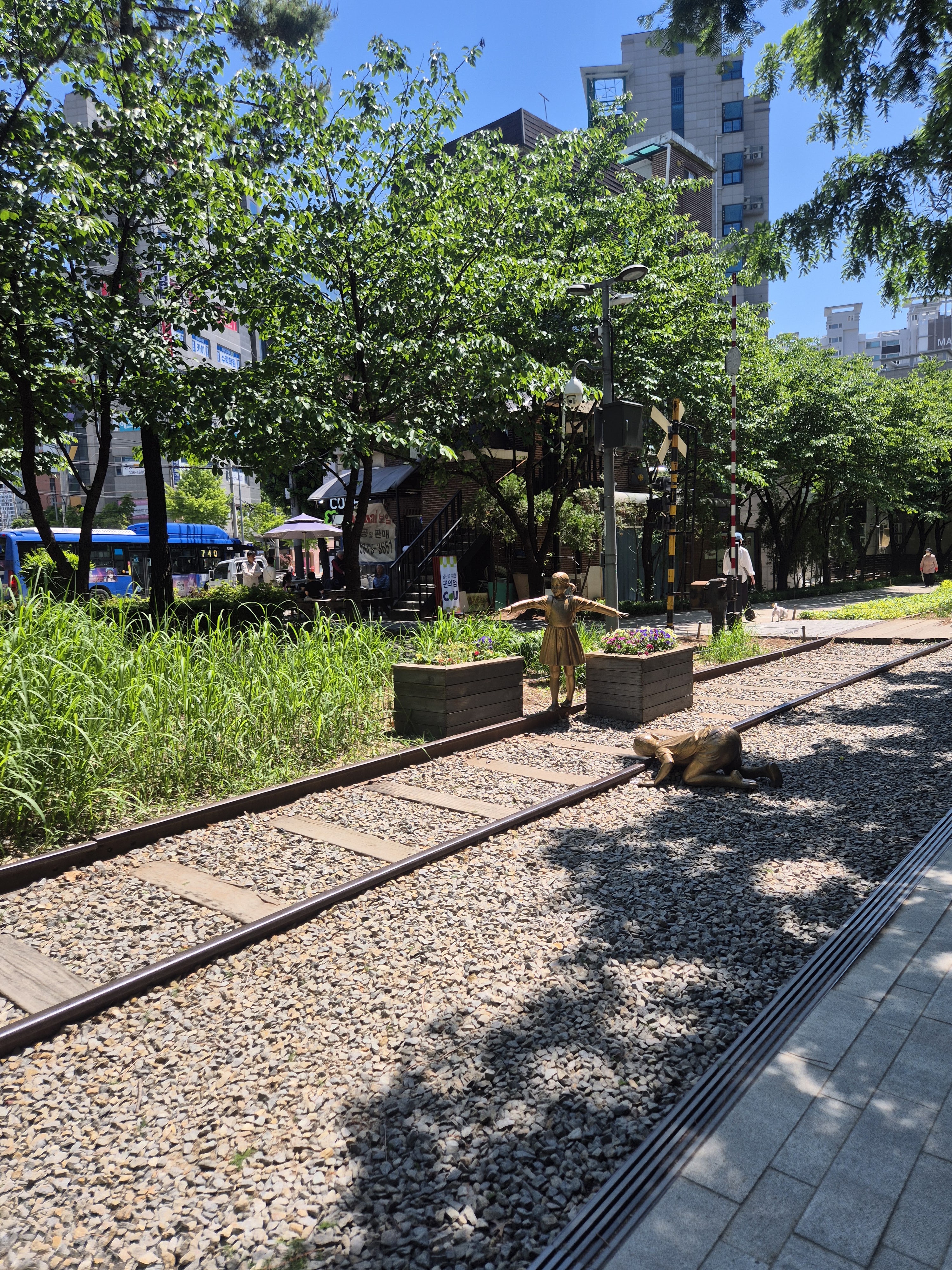A pathway beside train tracks surrounded by greenery, featuring two sculptures: one of a child standing and another of a child lying on the tracks. Urban buildings are visible in the background under a clear blue sky.